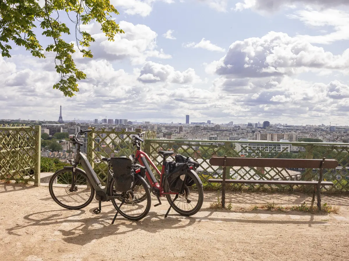 Two bicycles and a bench on the terrace of the Saint-Cloud park, facing the rooftops of Paris and the Eiffel Tower under a cloudy sky