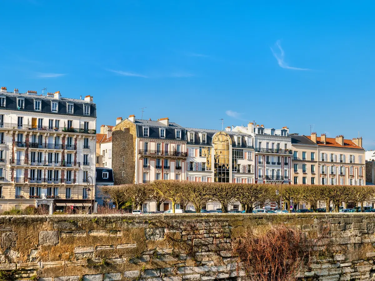 The apartment buildings of Vincennes and their balconies under a blue sky, above a stone wall and a row of pruned trees