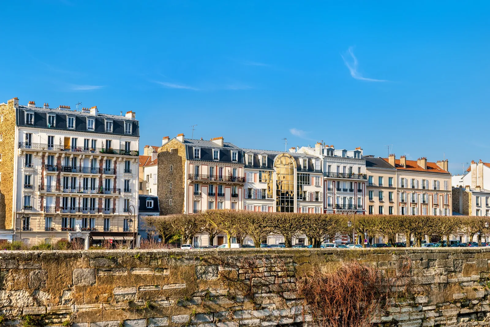 The apartment buildings of Vincennes and their balconies under a blue sky, above a stone wall and a row of pruned trees
