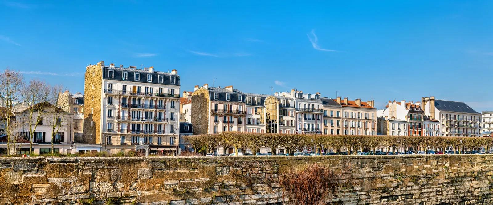 The apartment buildings of Vincennes and their balconies under a blue sky, above a stone wall and a row of pruned trees