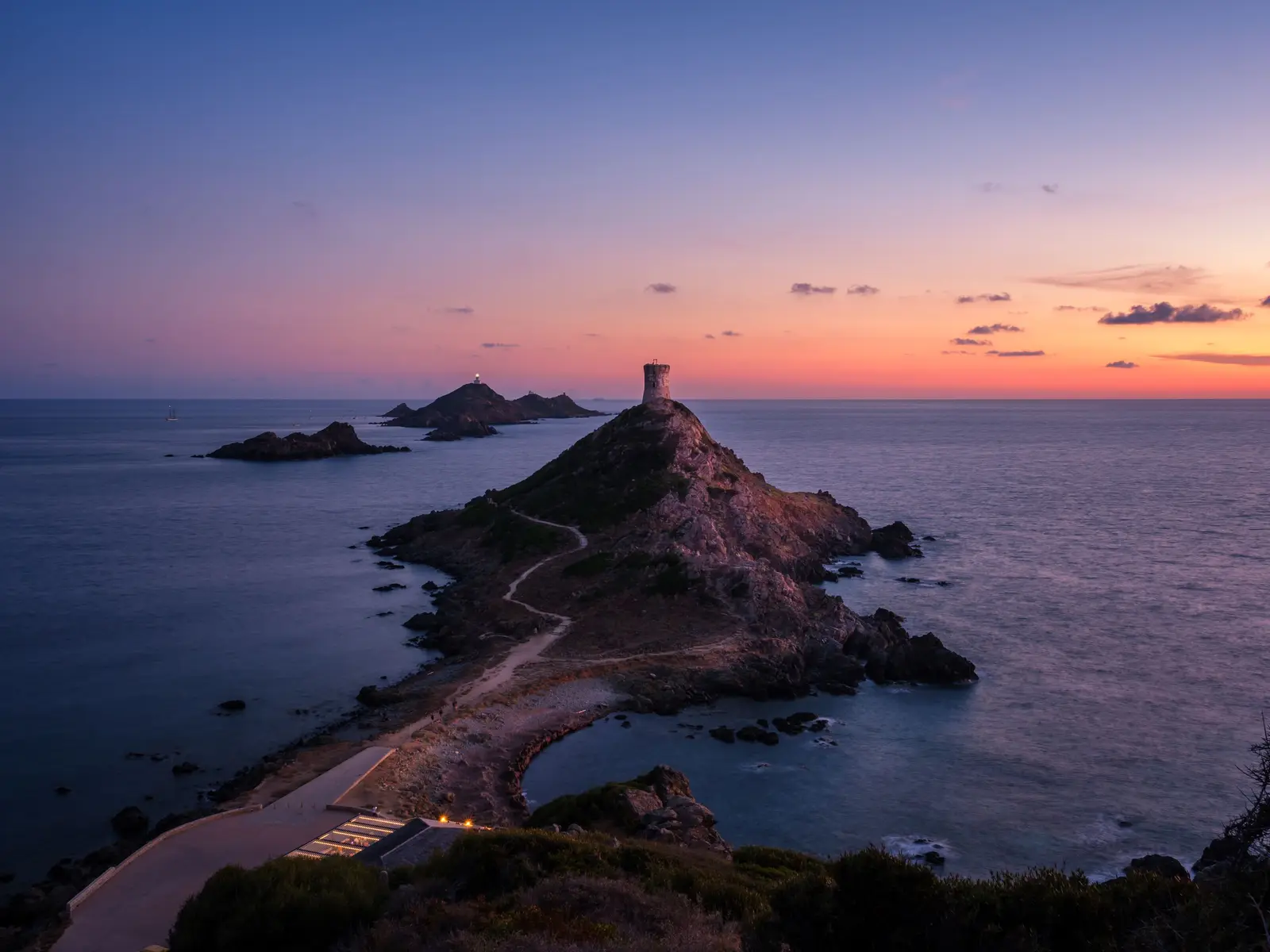 A torre genovesa da ponta da Parata ao crepúsculo, o caminho que sobe até ela e, ao largo, as ilhas Sanguinárias e o seu farol sob um céu rosa e laranja