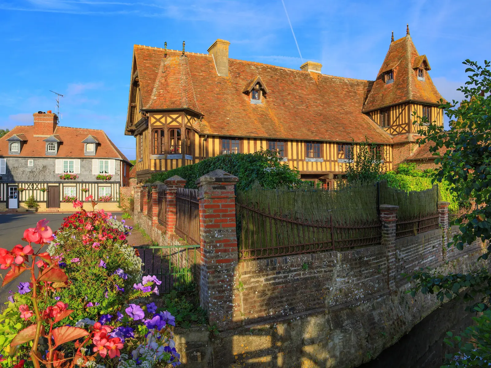 Le manoir à colombages de Beuvron-en-Auge, ses pans de bois sur fond ocre, son grand toit de tuiles et sa tourelle, derrière un mur de brique et des massifs de fleurs