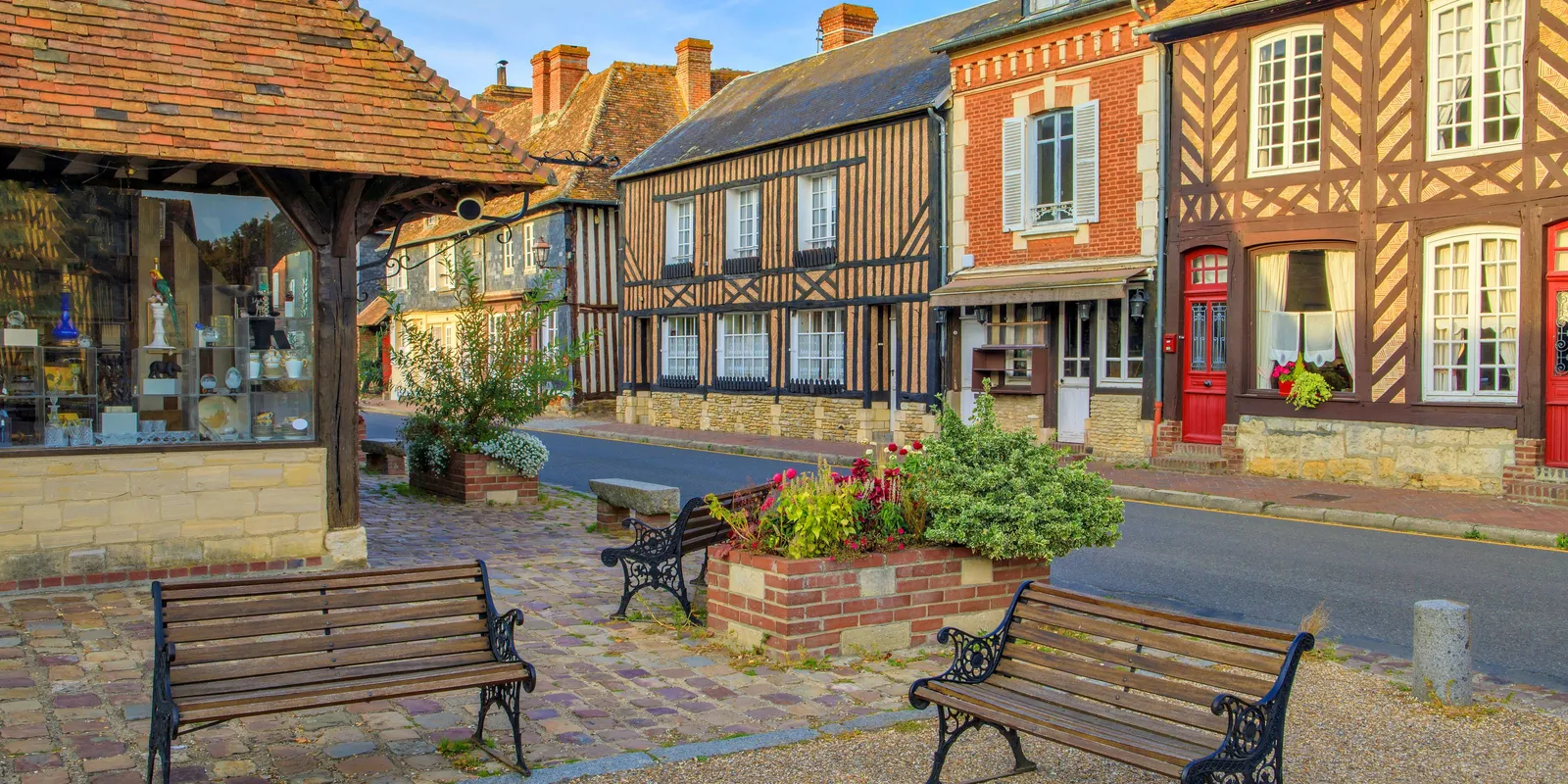 La place de Beuvron-en-Auge, ses bancs, ses jardinières fleuries et la halle ancienne, devant une rangée de maisons à colombages et une maison de brique aux portes rouges