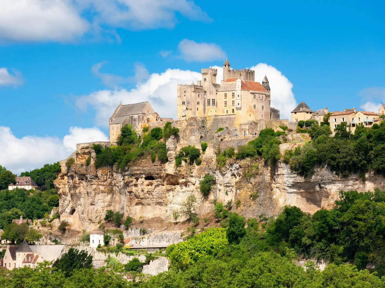 O castelo de Beynac no alto da sua falésia calcária, sobre as casas da aldeia e os bosques, sob um céu azul com grandes nuvens brancas