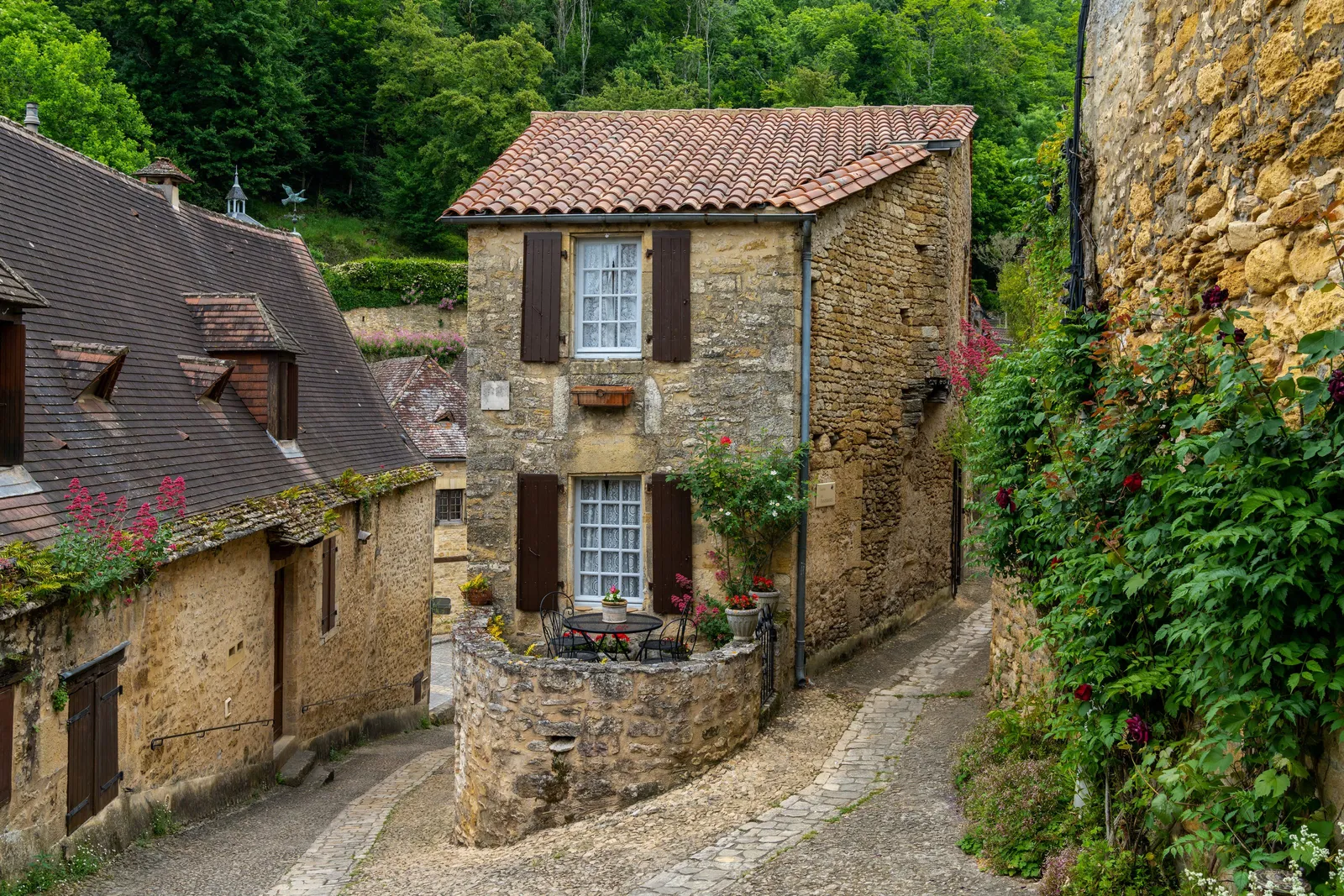 Uma pequena casa de pedra dourada de portadas castanhas entre duas ruelas calcetadas de Beynac-et-Cazenac, com o seu terraço redondo florido de gerânios e roseiras trepadeiras