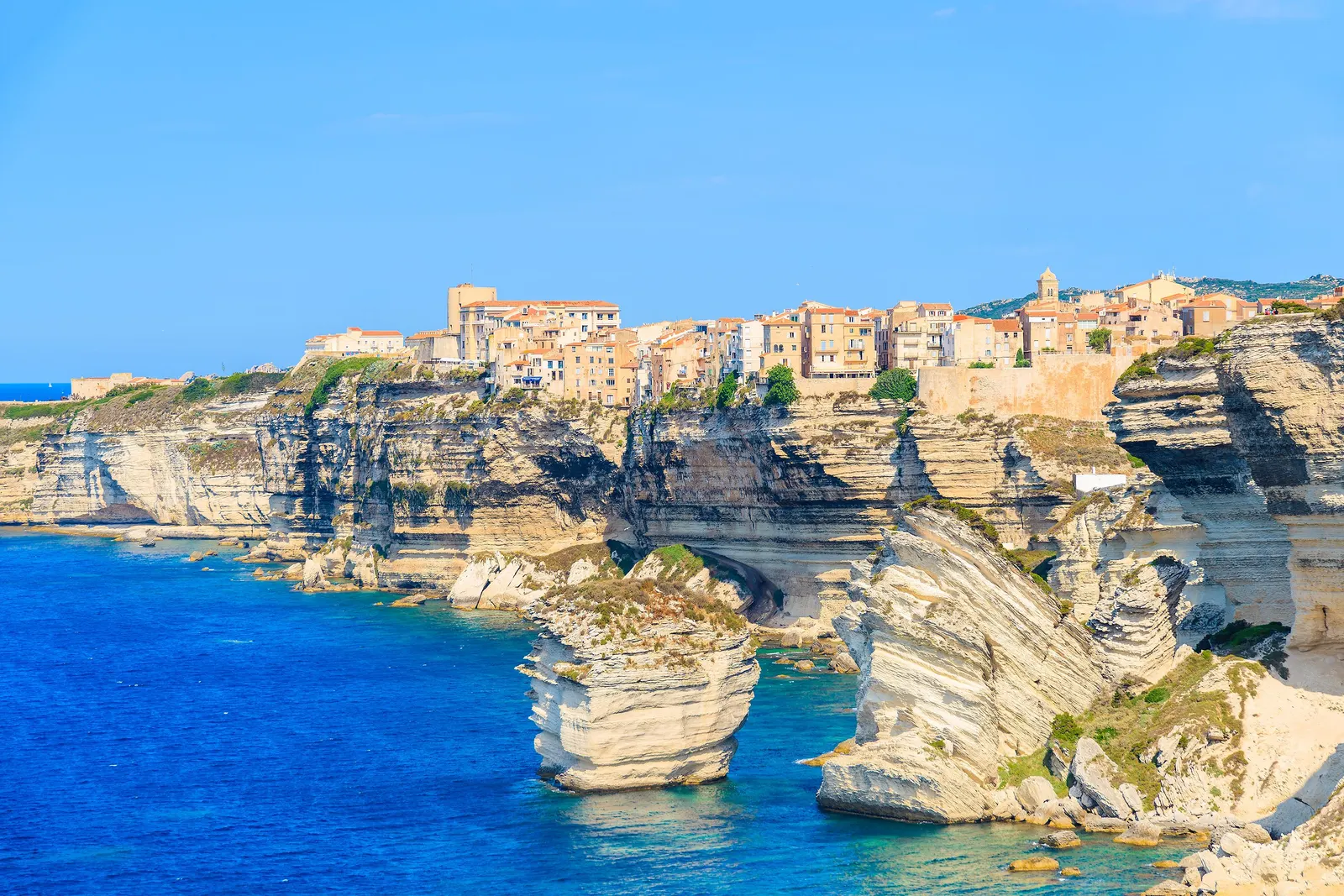 La haute ville de Bonifacio perchée au bord des falaises calcaires blanches, au-dessus d’une mer d’un bleu profond, avec le rocher du Grain de sable détaché de la côte
