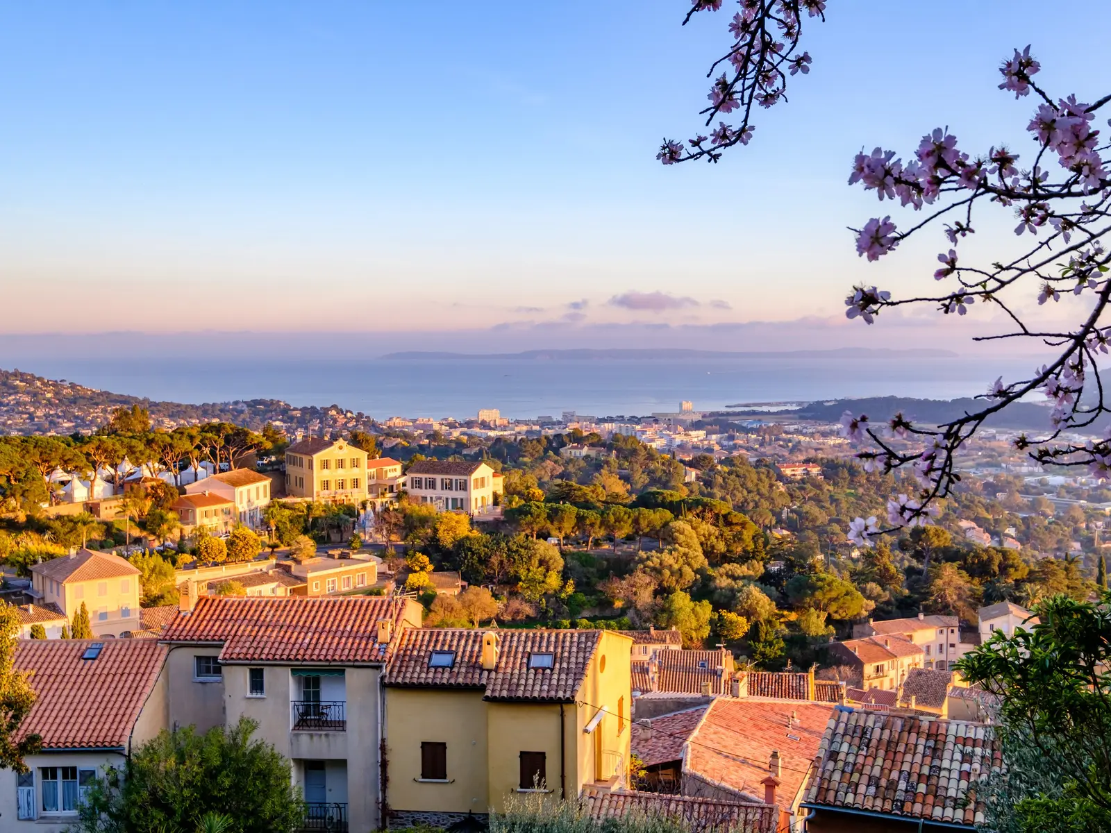 A vista do alto da aldeia de Bormes-les-Mimosas sobre os telhados e o mar, ramos de amendoeira em flor em primeiro plano