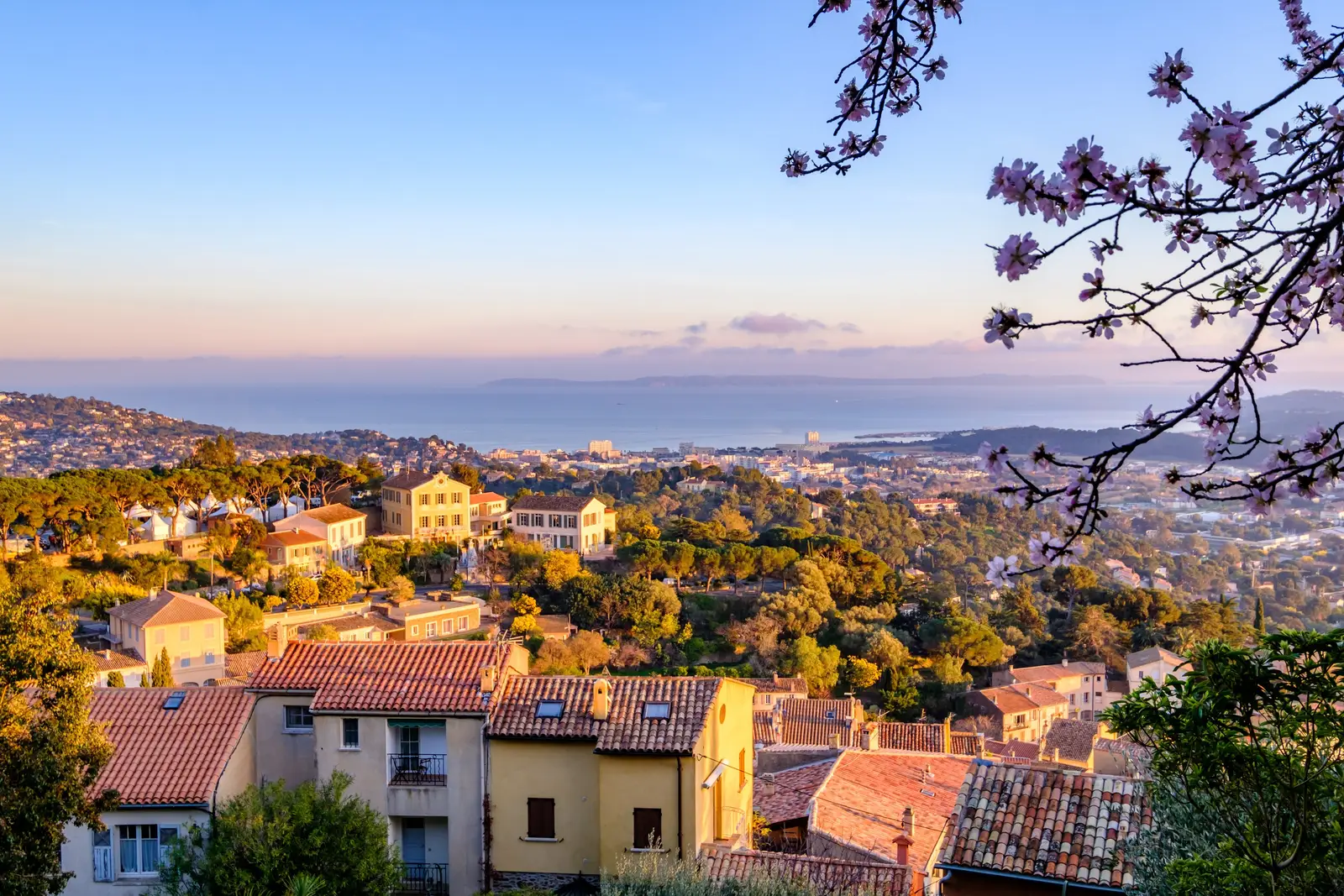 A vista do alto da aldeia de Bormes-les-Mimosas sobre os telhados e o mar, ramos de amendoeira em flor em primeiro plano