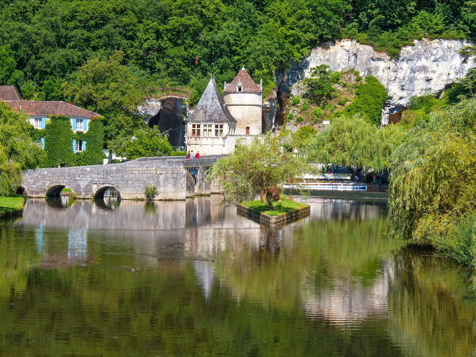 A ponte de pedra em cotovelo sobre o rio Dronne em Brantôme, o pavilhão renascentista com a sua torre redonda e a falésia calcária branca, com salgueiros-chorões refletidos na água calma