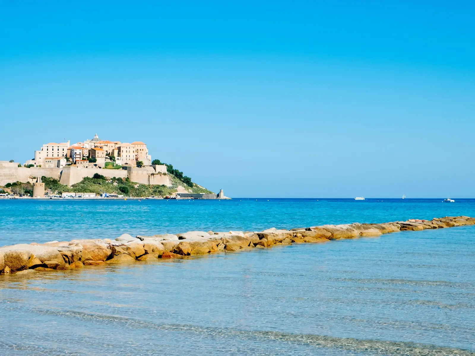 La citadelle de Calvi sur son promontoire, vue depuis la plage, au-delà d’une eau turquoise transparente et d’une jetée de rochers, sous un grand ciel bleu
