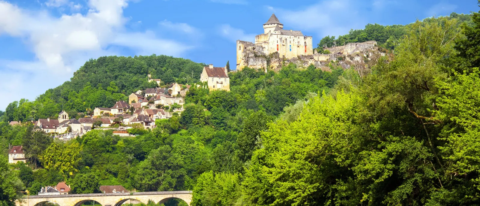 O castelo de Castelnaud no seu esporão arborizado, a aldeia em socalcos na encosta e a ponte de pedra sobre o rio Dordonha, com o rio em primeiro plano