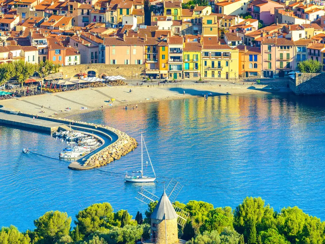 Collioure vue du ciel, ses maisons colorées, sa plage et son port, un moulin dans les pins au premier plan