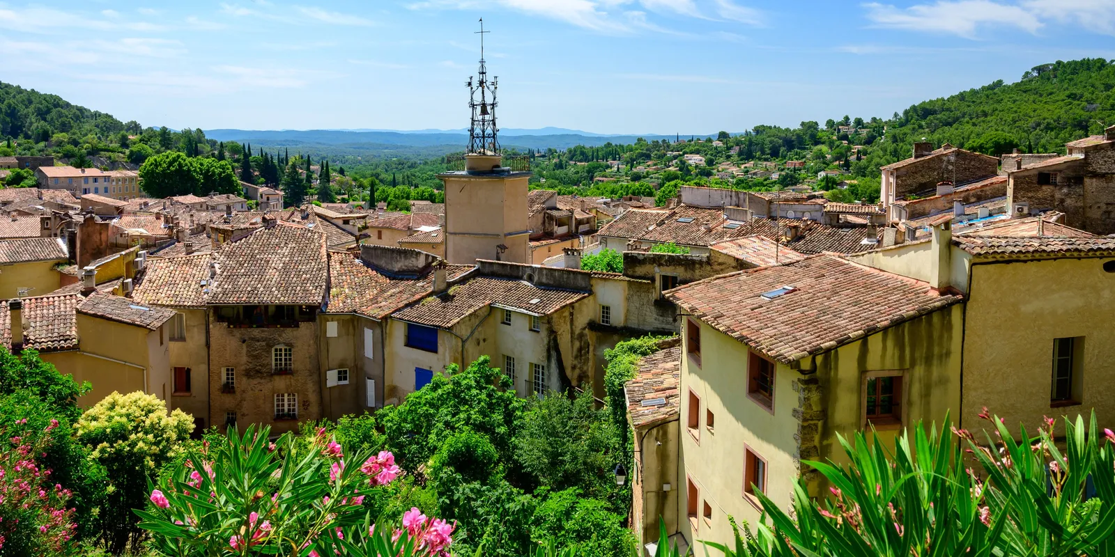 Les toits de tuiles de Cotignac autour d’une tour coiffée d’un campanile en fer forgé, avec des lauriers-roses en fleurs au premier plan et des collines boisées au loin