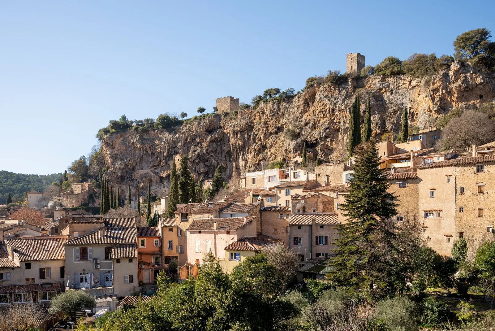 Les maisons ocre de Cotignac serrées au pied de la grande falaise de tuf, couronnée de deux tours médiévales, entre les cyprès, sous un ciel bleu