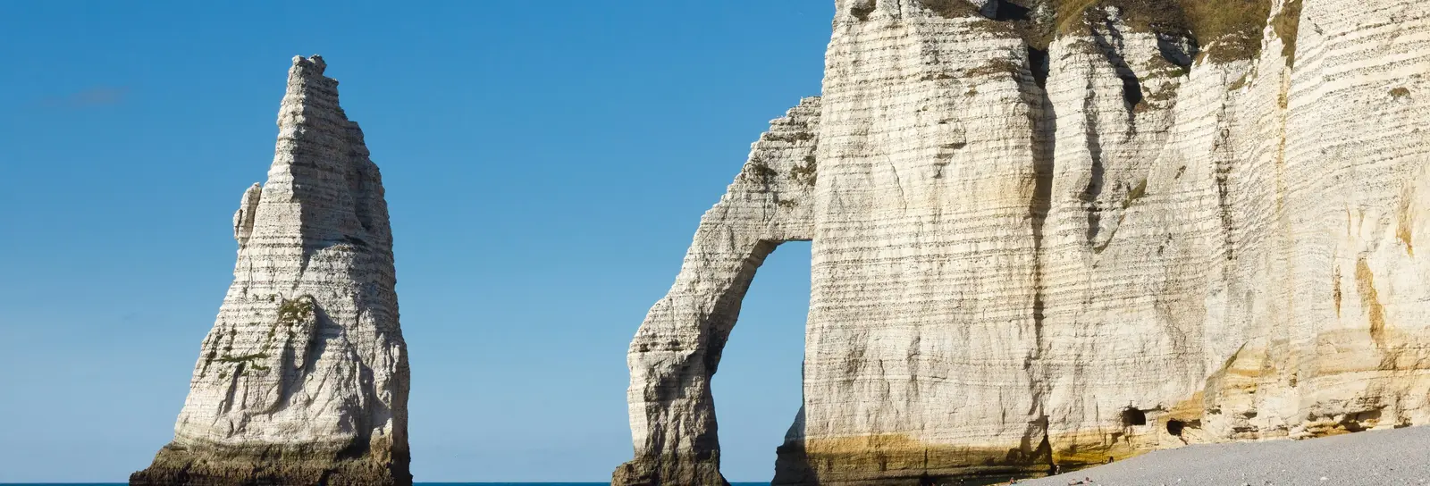 The Aiguille and the Porte d’Aval arch in Étretat seen from the shingle beach, under a blue sky