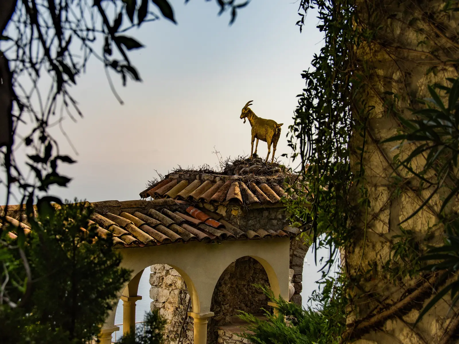 A estátua da cabra de ouro no telhado de uma loggia de Èze village, entre os ramos, em contraluz