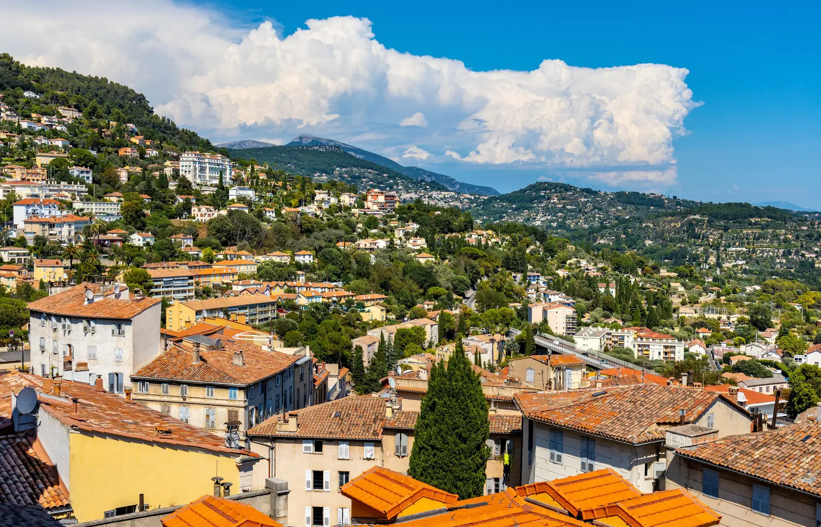 As colinas de Grasse e as suas villas no meio do verde, acima dos telhados de telha da cidade, os Pré-Alpes ao fundo