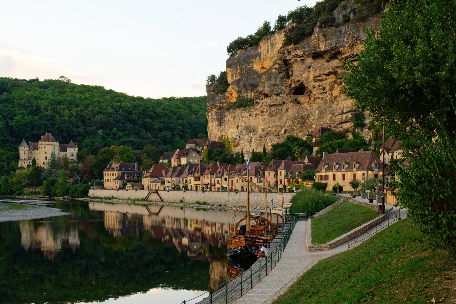 As casas de pedra dourada de La Roque-Gageac alinhadas ao pé da falésia, à beira do rio Dordonha, com as gabarras atracadas no cais e o reflexo da aldeia na água calma do fim de tarde