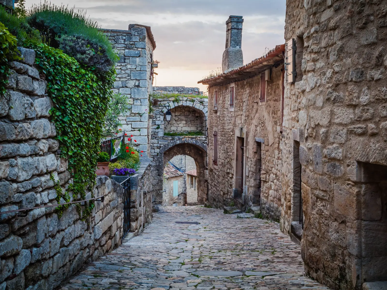 Uma ruela calcetada de Lacoste entre casas de pedra, que conduz a um arco duplo, com hera e flores no muro, na luz suave do fim de tarde