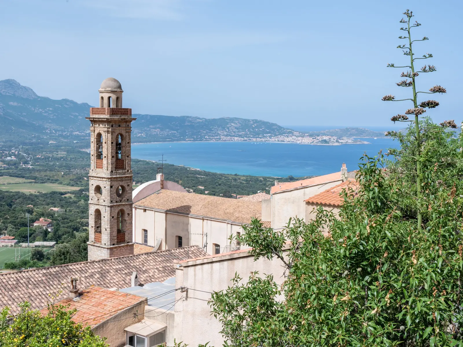 O campanário de pedra da igreja de Lumio e os telhados desta aldeia da Balagne, com o golfo de Calvi turquesa, a cidade de Calvi e as montanhas ao longe