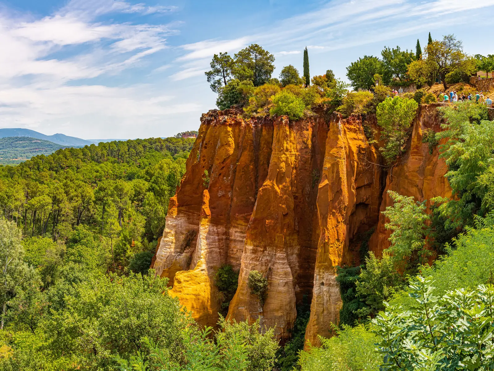 As falésias de ocre laranja e amarelo de Roussillon, coroadas de pinheiros e ciprestes, no meio de um pinhal verde sob um céu azul velado