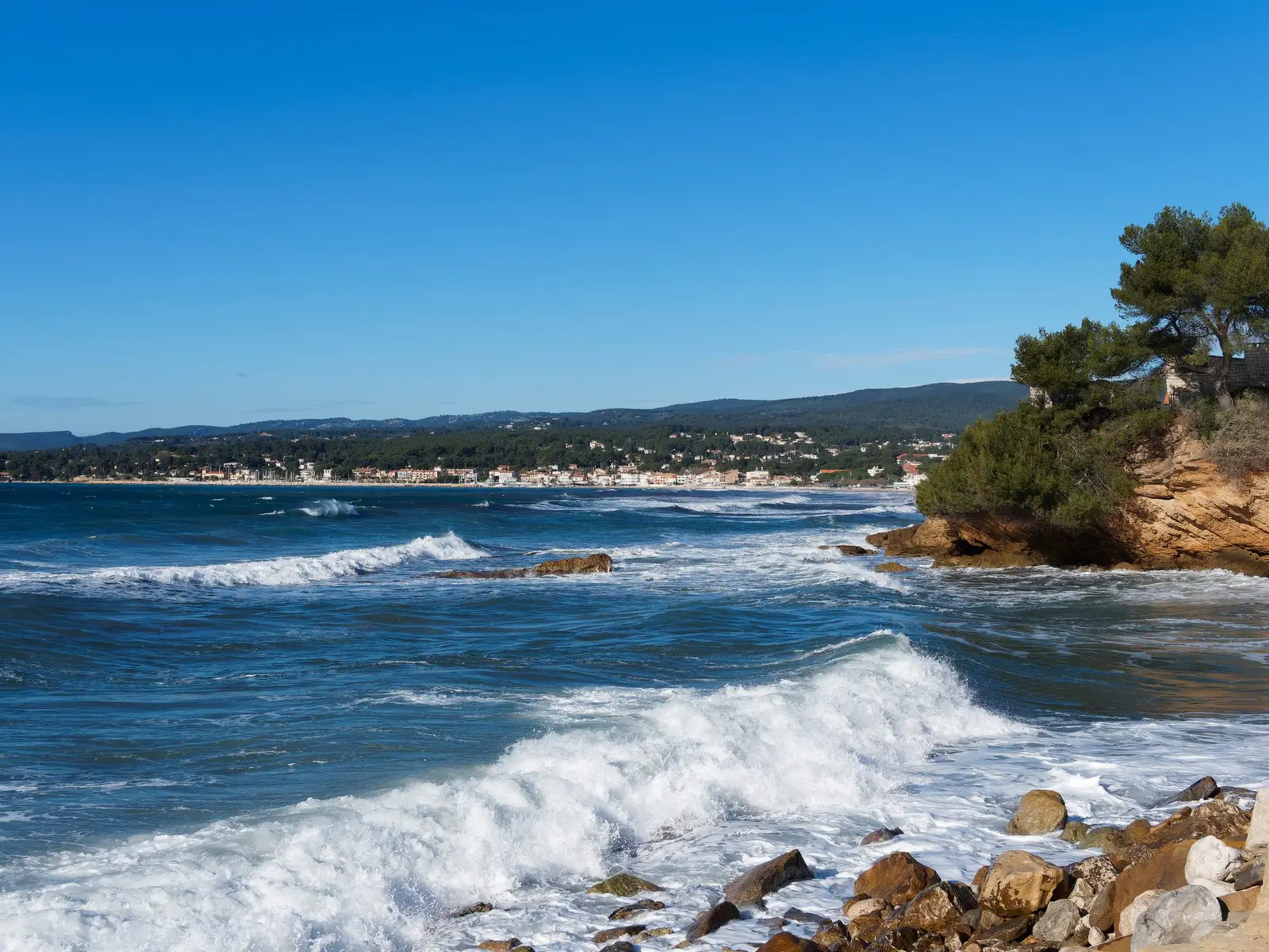 A baía de Les Lecques em Saint-Cyr-sur-Mer, ondas sobre as rochas, pinheiros e as casas da costa ao longe