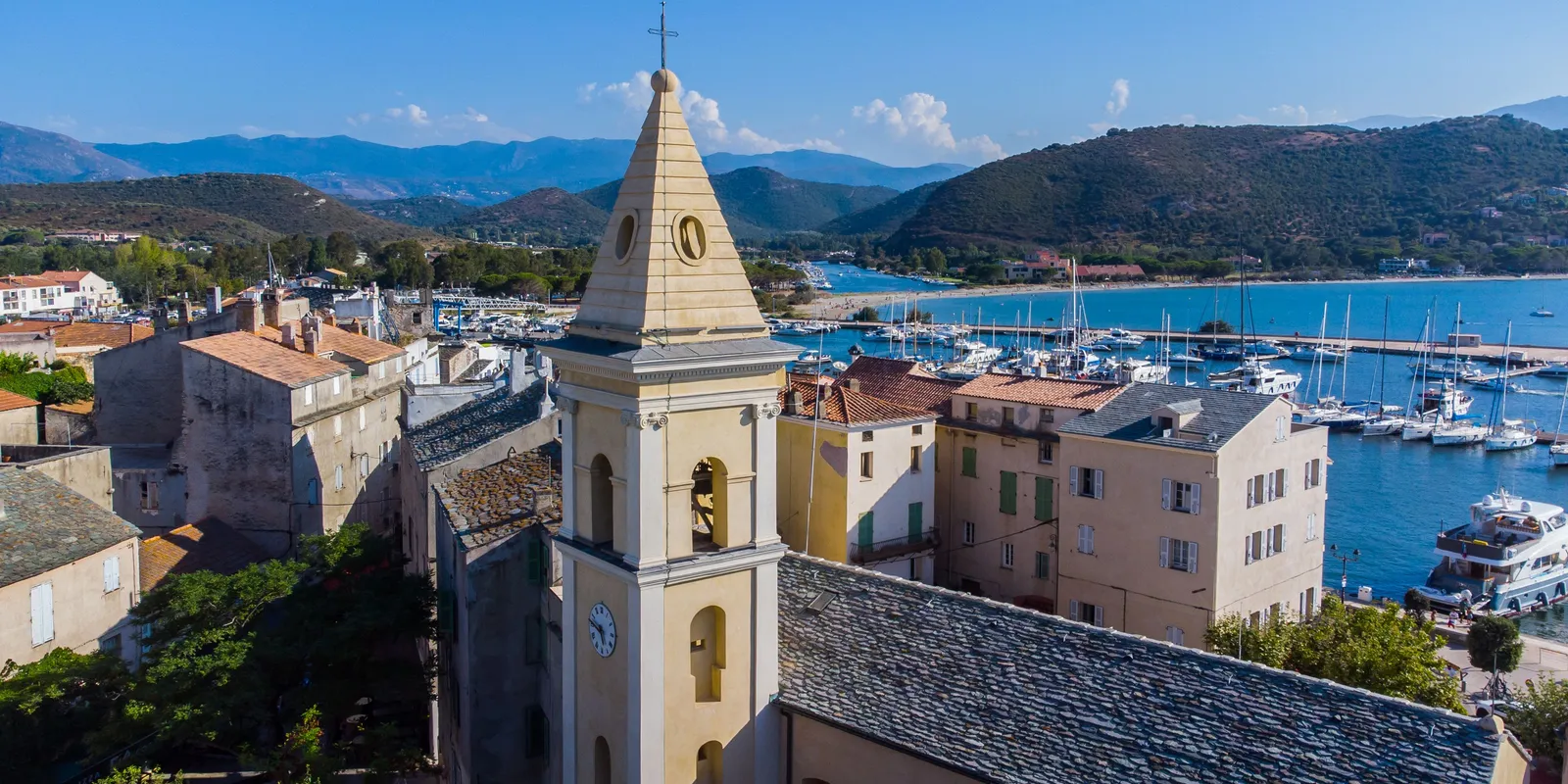 Saint-Florent vista do céu, com o campanário com relógio da igreja, os telhados de lousa da cidade velha, a marina cheia de veleiros e o golfo rodeado de colinas