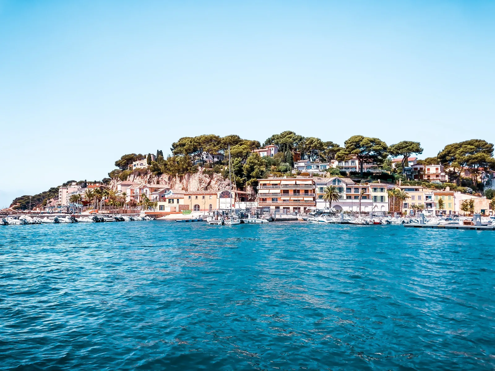 Les bateaux amarrés au port de Sanary-sur-Mer, les façades colorées et les villas sous les pins, vus depuis la mer