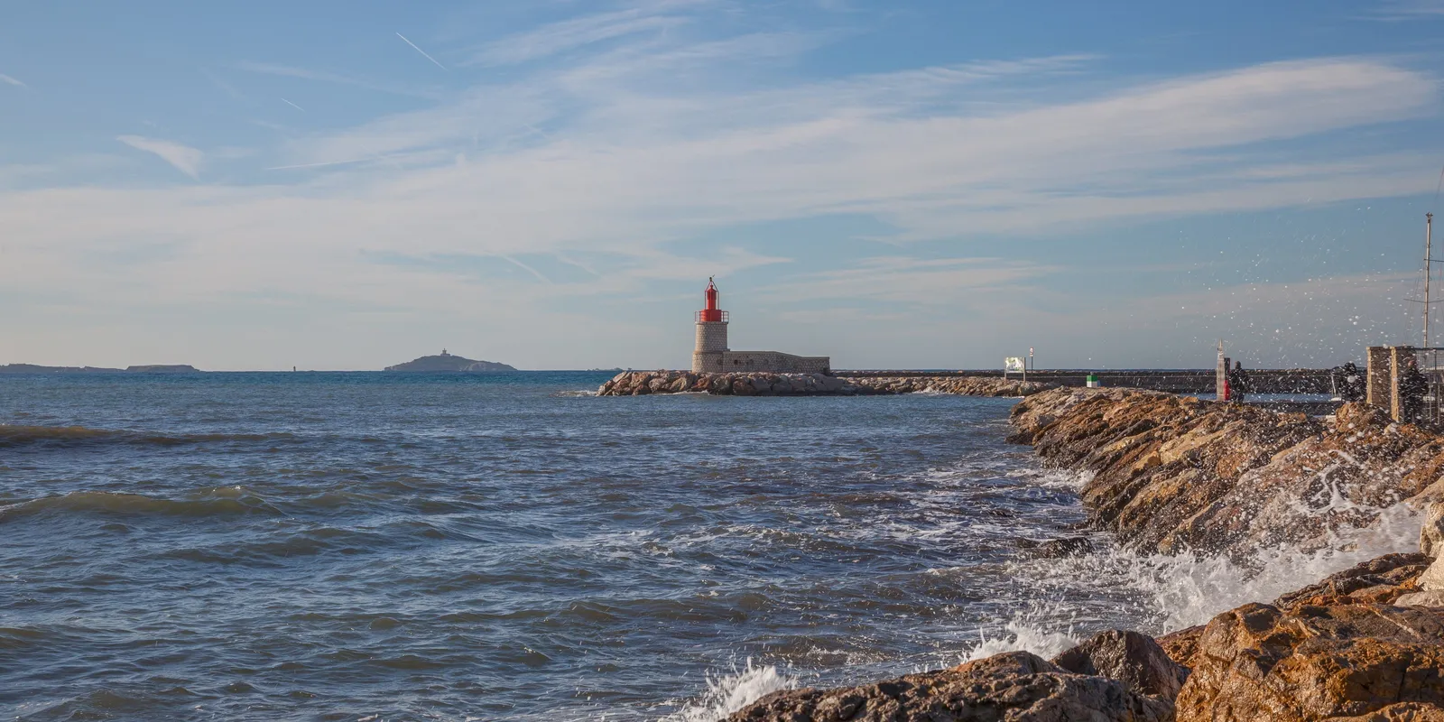 O farol vermelho de Sanary-sur-Mer no extremo do molhe, as ondas nos rochedos e as ilhas ao largo