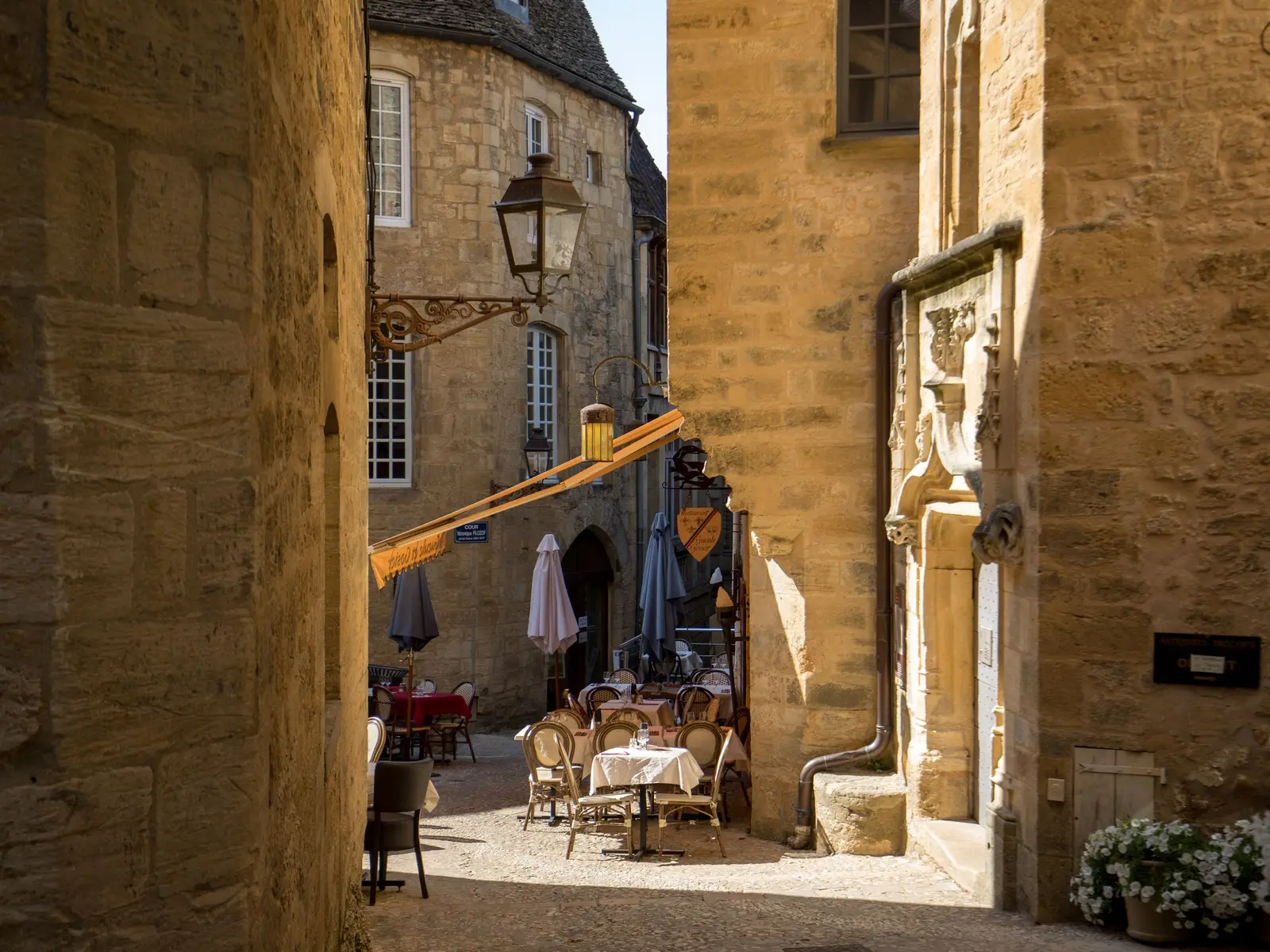 Uma ruela de pedra dourada da cidade velha de Sarlat-la-Canéda, com uma esplanada de restaurante posta, uma lanterna de ferro forjado e um portal de pedra esculpida