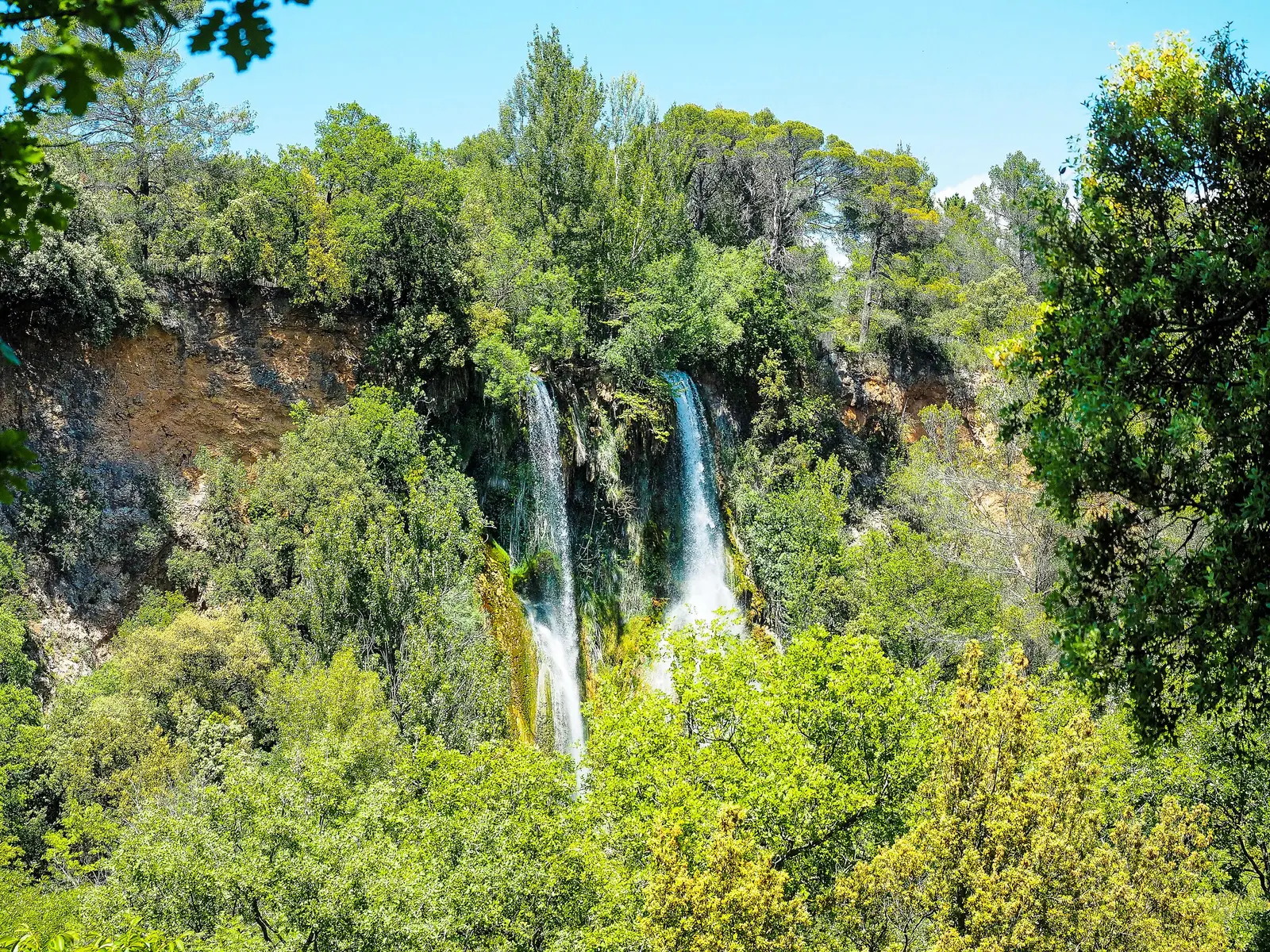 La cascade de Sillans tombant en deux chutes d’une falaise ocre au milieu d’une végétation dense, sous un ciel bleu