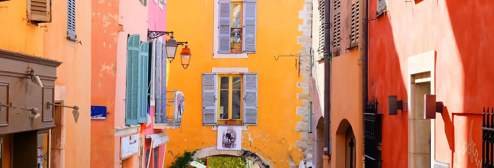 A lane in old Valbonne with its ochre, pink and orange façades, its blue shutters and a stone arch at the end of the street
