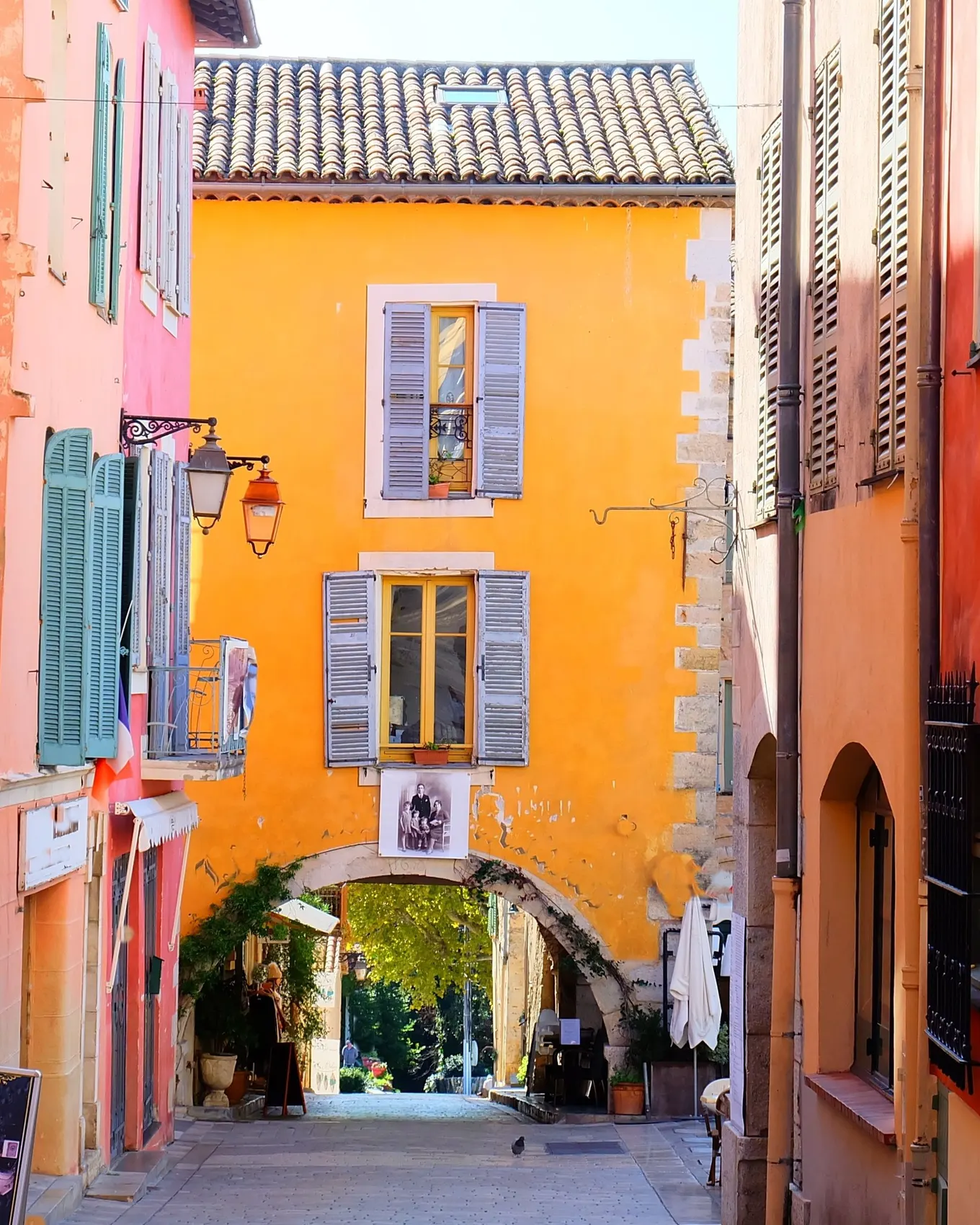 A lane in old Valbonne with its ochre, pink and orange façades, its blue shutters and a stone arch at the end of the street