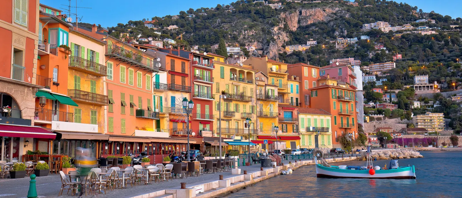 O cais de Villefranche-sur-Mer ao amanhecer, as suas fachadas coloridas, esplanadas e um barco à beira da água