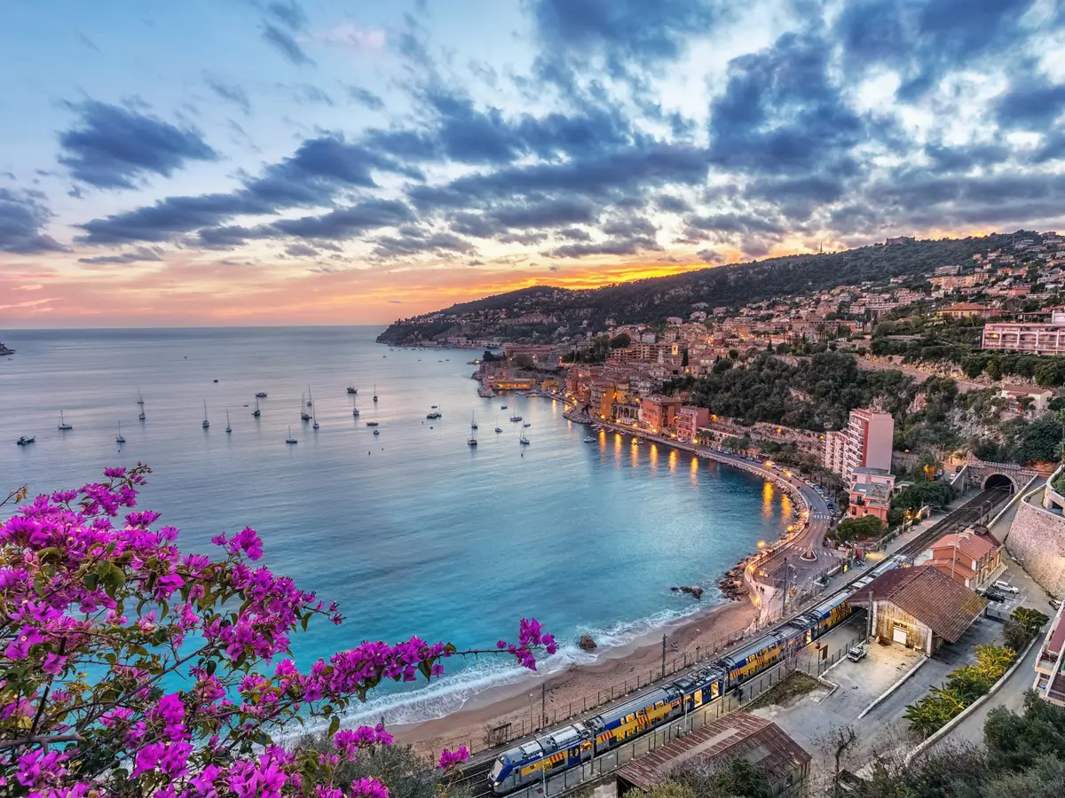 La rade de Villefranche-sur-Mer et ses maisons au coucher du soleil, vues des hauteurs à travers les bougainvilliers en fleurs